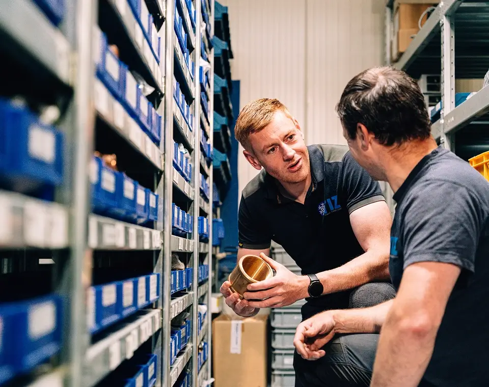 Twee collega's overleggen in het magazijn over een koperen fitting, tussen de stellingen met onderdelenbakken