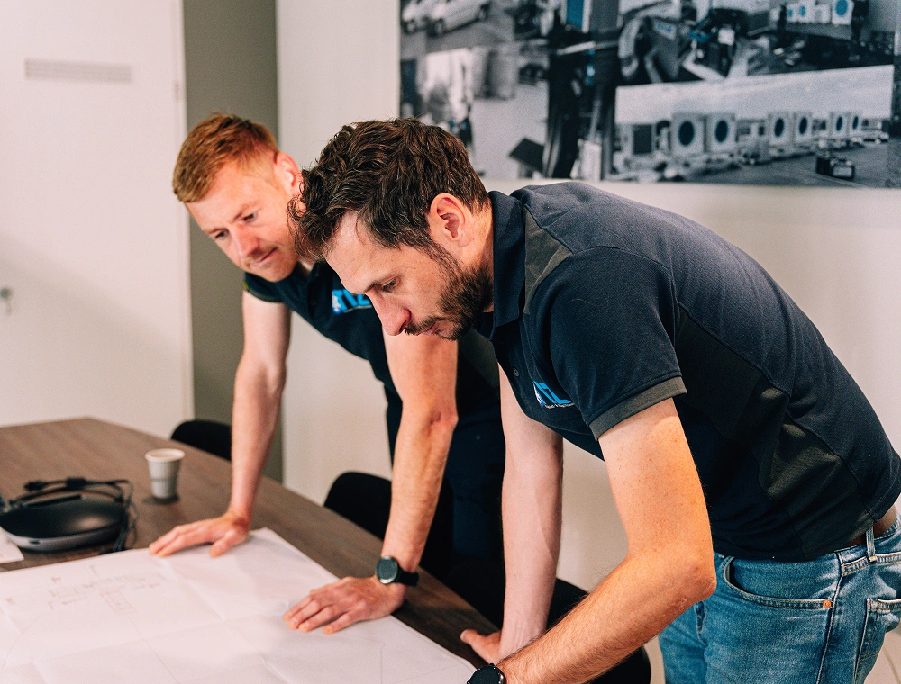 Twee collega's buigen zich over een bouwtekening op de vergadertafel om het project door te nemen