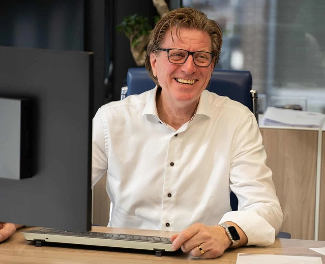 Directeur lachend achter zijn bureau met beeldscherm en toetsenbord op kantoor bij TIZ