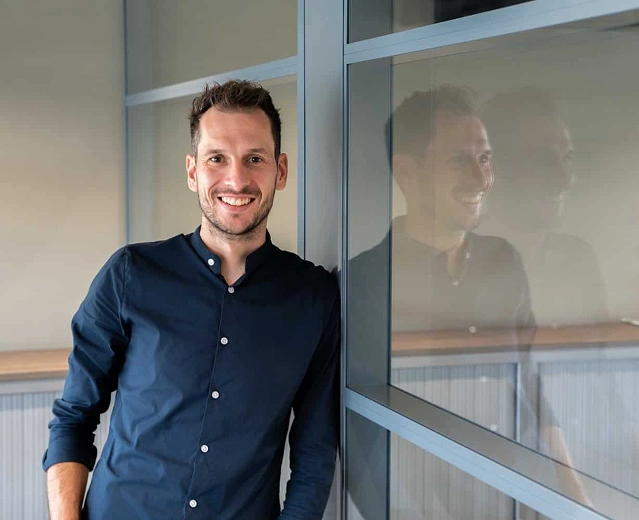 Medewerker in donker overhemd leunt lachend tegen een glazen scheidingswand op kantoor