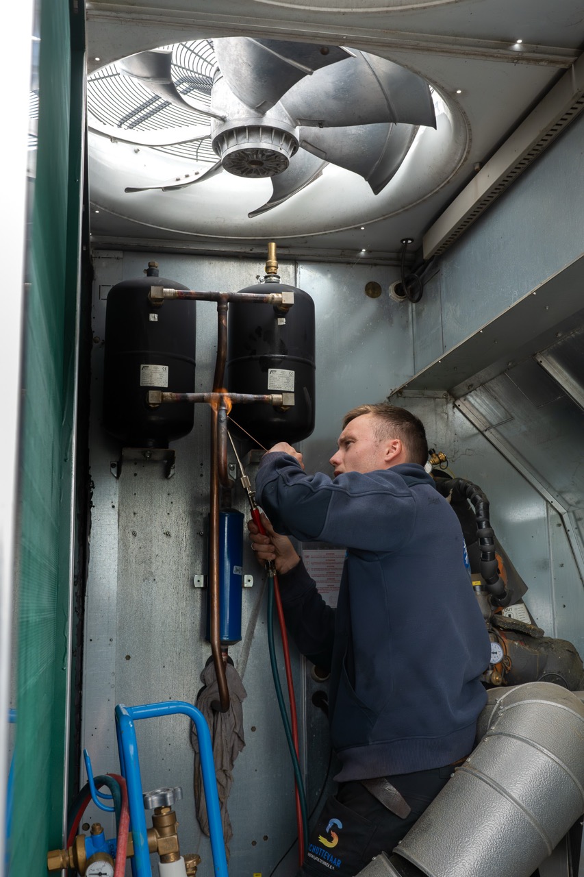 Monteur van TIZ soldeert koperen leidingen binnen een grote koelinstallatie tijdens reparatiewerkzaamheden.